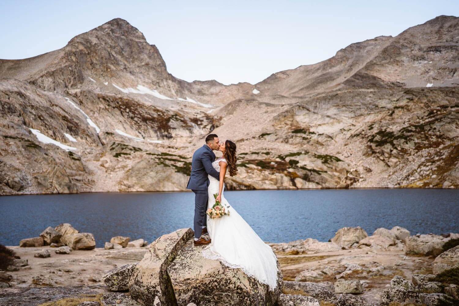 Bride and groom kissing on a rock at alpine lake in Colorado.