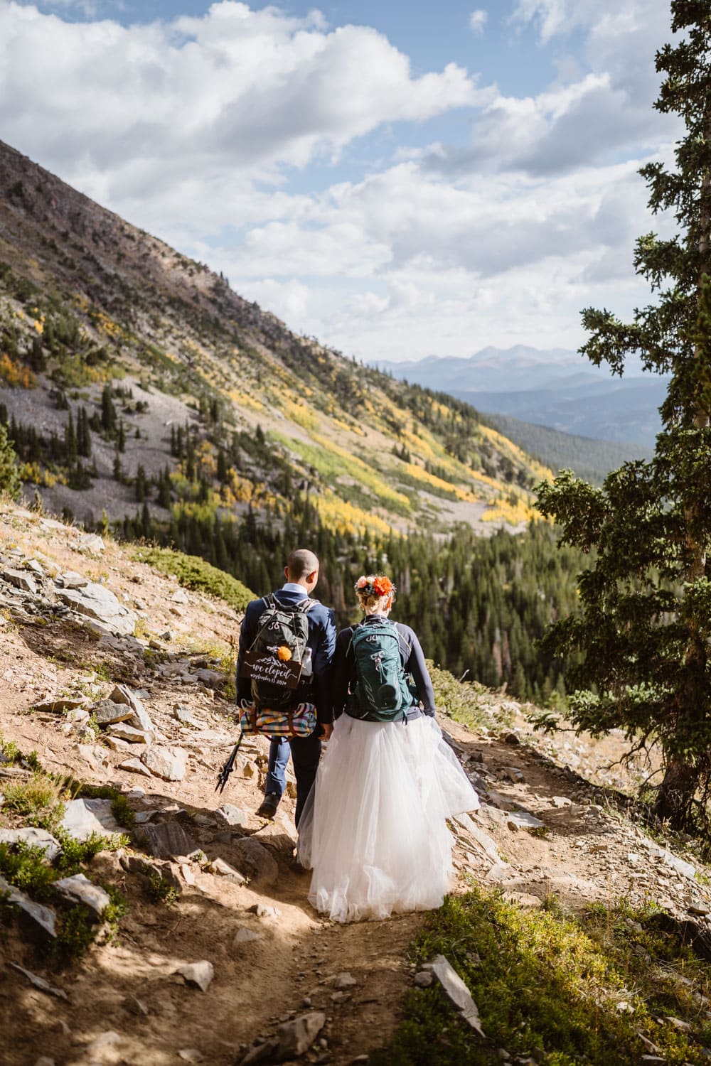 Bride and groom hiking down in their wedding attire in the mountains of Colorado.,