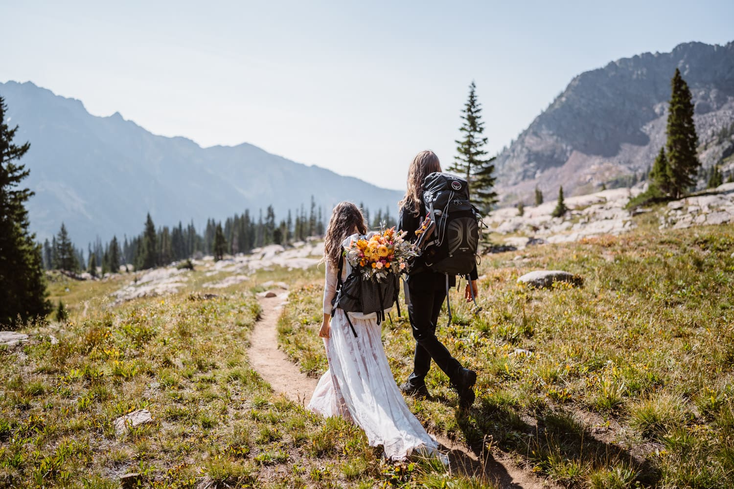 Bride and groom hiking away in their wedding clothes with backpacks,