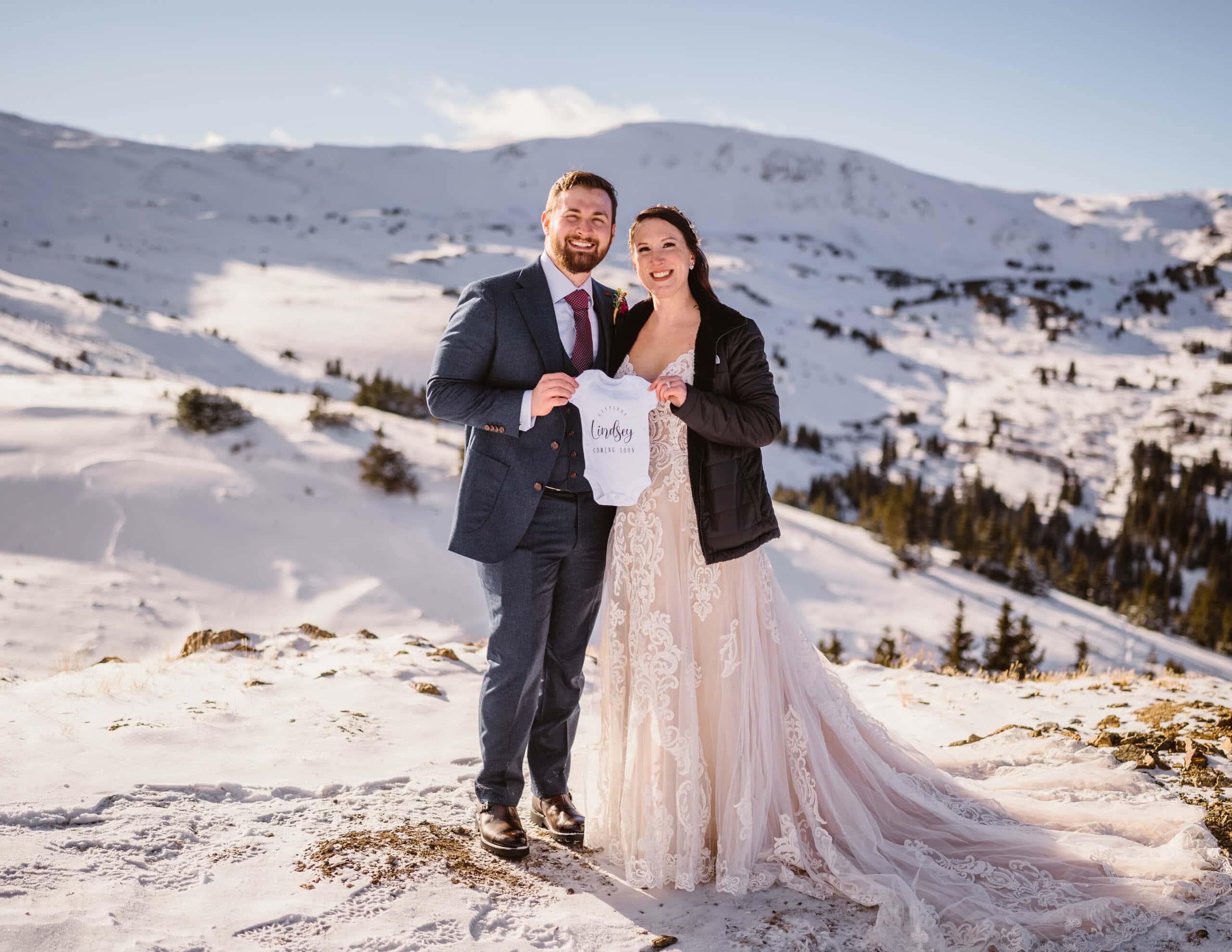 Bride and groom showing off their baby's outfit in the snow of Colorado.